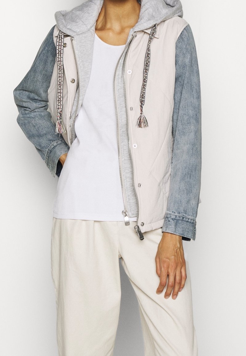 Letvægtsjakke med grå hætte, beige front og blå denimærmer, med dekorative frynser og en sølv lynlås.