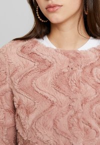 Růžový chlupatý svetr s vlnitým vzorem a měkkým plyšovým povrchem. V pozadí pár kulatých kovových kruhových náušnic.