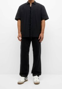 Hombre vestido con una camisa de botones negra de manga corta y textura, pantalones negros de pierna ancha y zapatillas blancas con rayas negras.