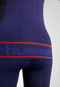 Gros plan d’une personne portant un legging de sport bleu foncé et un haut avec une ceinture rayée rouge et bleue affichant en partie le logo "hummel".