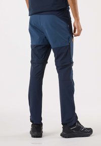 Marinblå andningsbara byxor med en kontrasterande ljusblå panel, elastisk midjeband och sidoförsedda blixtlås, kombinerade med svart robusta skor.