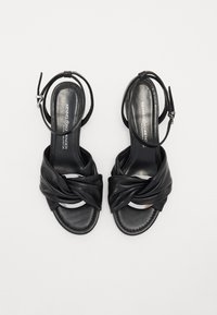 Sandales à talons hauts en cuir noir avec un design noué à l'avant, bout ouvert, sangle de cheville et accents argentés brillants. Texture lisse.