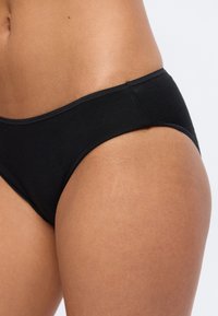 Κοντινό πλάνο ενός ατόμου που φορά μαύρο βαμβακερό εσώρουχο, εμφανίζοντας το ισχίο και την κάτω κοιλιακή χώρα μπροστά σε απλό ανοιχτό φόντο.