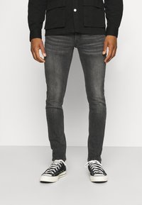 Jeans gris de corte ajustado hechos de denim, combinados con zapatillas de lona negras con cordones blancos y suelas de goma sobre un fondo liso.