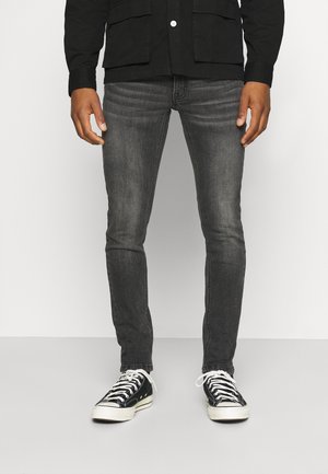 Jeans gris de corte ajustado hechos de denim, combinados con zapatillas de lona negras con cordones blancos y suelas de goma sobre un fondo liso.