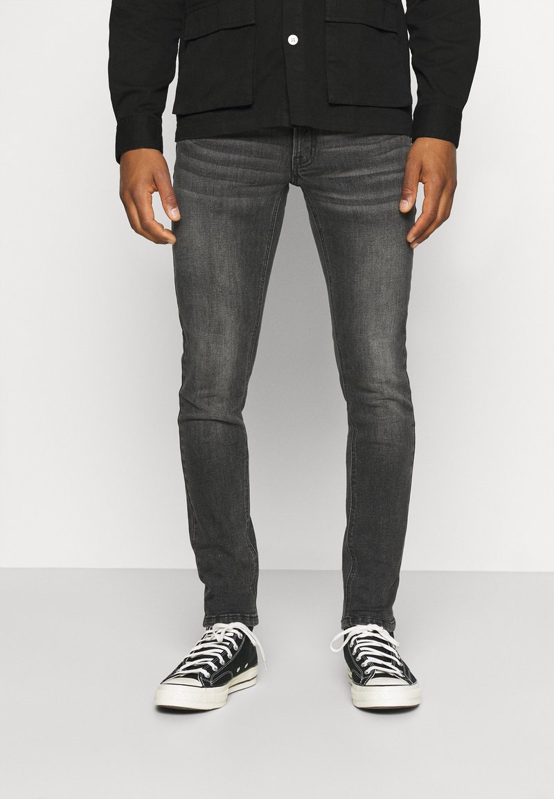 Jeans gris de corte ajustado hechos de denim, combinados con zapatillas de lona negras con cordones blancos y suelas de goma sobre un fondo liso.