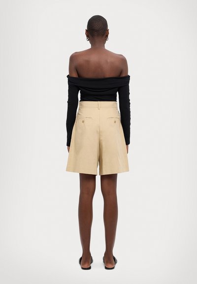 Schwarzes Off-Shoulder-Top, hellbeige, hochgeschnittene Shorts, zwei Gesäßtaschen mit Knöpfen, kombiniert mit schwarzen flachen Sandalen.
