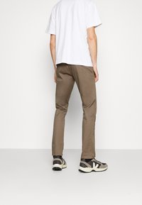 Pantalon marron coupe slim, en mélange de coton. Associé à un t-shirt blanc uni et des baskets de ton neutre, debout sur un fond blanc.