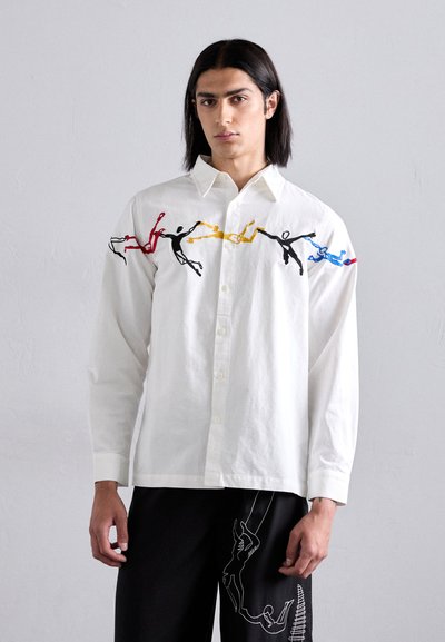 Chemise blanche à manches longues avec fermeture à boutons, ornée de figures brodées colorées en rouge, jaune, bleu et noir le long des épaules.