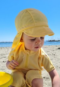 Gul solhat og kortærmet dragt, lavet af let materiale, med lynlås foran, på et barn, der sidder på en sandstrand nær vandet.