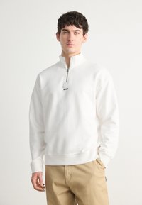 Hvit zip-neck sweatshirt med subtil elastisk linning og mansjetter, med en liten svart "HUGO"-logo på brystet. Standard passform.
