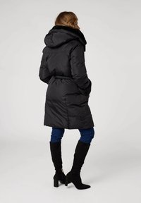 Manteau en duvet noir avec une capuche doublée de fourrure, une taille ceinturée et des poches latérales. Porté sur un jean bleu et des bottes noires jusqu'au genou.