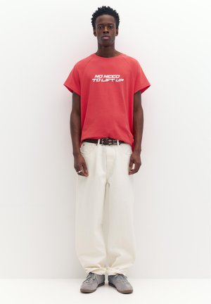 Junger Mann steht, trägt ein rotes T-Shirt mit dem Aufdruck „NO NEED TO LIFT UP“, weiße lockere Hose, schwarzen Gürtel und graue Sneakers, vor einfachem Hintergrund.