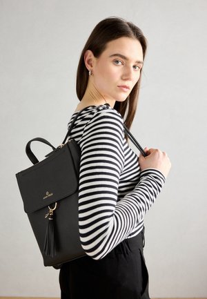 Vrouw draagt een zwart-wit gestreept shirt met lange mouwen en een zwarte Anna Field rugzak met een tassel-detail op haar schouder.