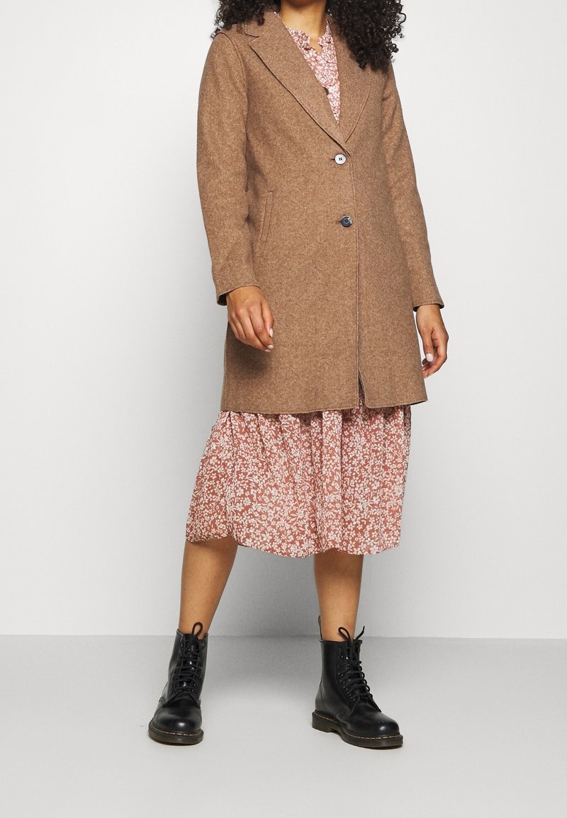 Femme portant un manteau en laine marron sur une robe fleurie rose et des bottines noires à lacets, debout devant un fond blanc uni.