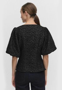 Kvinna med brunt hår i hästsvans bär en svart texturerad blommig blus med korta ärmar och mörka jeans, vänd bort från kameran mot en enkel bakgrund.
