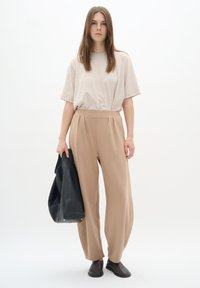 Beige t-shirt med korta ärmar, tillsammans med vida beige byxor. Modellen håller en svart läderväska. Bruna ankelstövlar bärs.