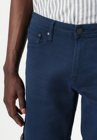 Jeans bleu marine foncé en denim avec une texture lisse. Comprend une poche avant, un bouton en métal et des passants de ceinture. Aucun motif visible.