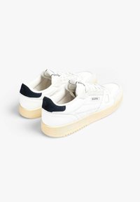 Zapatillas de lona blancas con un acento en el talón de color azul marino y suelas de goma crema. Cuentan con un clásico cierre de cordones y un sutil detalle de logo en el lateral.