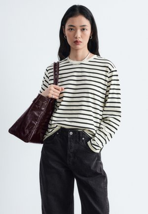 Femme portant un chemisier à manches longues rayé noir et blanc et un pantalon noir, portant un grand sac en cuir marron foncé sur l'épaule.