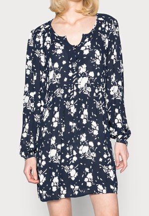 Kvinde iført navyblå langærmet kjole med hvidt blomsterprint og rund halsudskæring med hak, stående mod en lys baggrund.