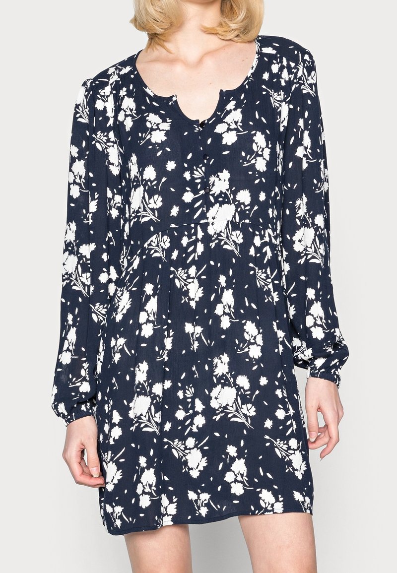 Femme portant une robe bleu marine à manches longues avec motif floral blanc et encolure ronde avec une encoche, debout sur un fond clair.