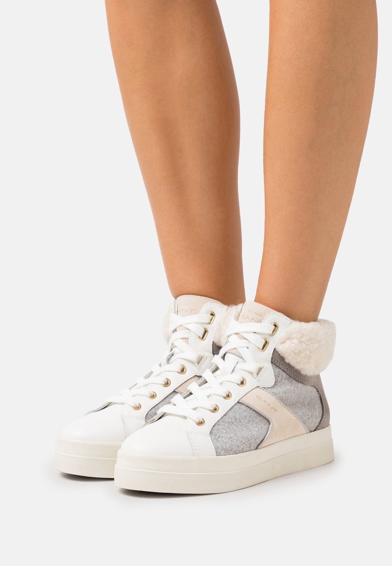 Ben som har på sig vita, beige och grå höga sneakers med foder av päls och vita skosnören, står på en enkel ljus bakgrund.