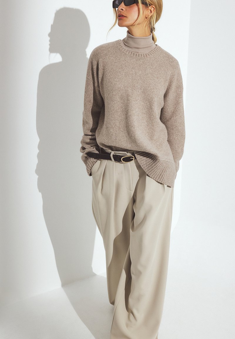 Helle beige Strickpullover mit einem gerippten Ausschnitt, kombiniert mit lockeren, cremefarbenen Hosen mit Plissee-Details und einem dunklen Gürtel.