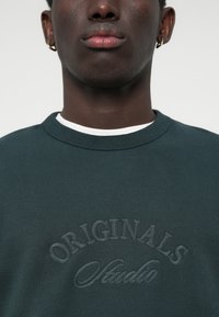 Tumedat rohelised dressipluusid, millel on ribatöötlusega ümar kaelus ja esipaneelil reljeefne "ORIGINALS Studio" tekst. Klassikaline, pehme kangakvaliteet.