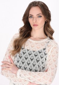 Top in pizzo bianco con motivo intricati, maniche lunghe e polsini ondulati sopra un vestito nero, evidenziando la texture e il design dettagliato.