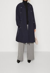 Parka imperméable bleu marine avec capuche ajustable par cordon de serrage, fermeture éclair à l'avant et poignets élastiques, associée à un pantalon gris à jambes larges et des chaussures noires.