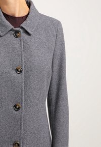 Kvinna som bär en grå ullkappa med fyra bruna knappar över en mörkbrun polotröja, avskuren vid halsen och midjan.