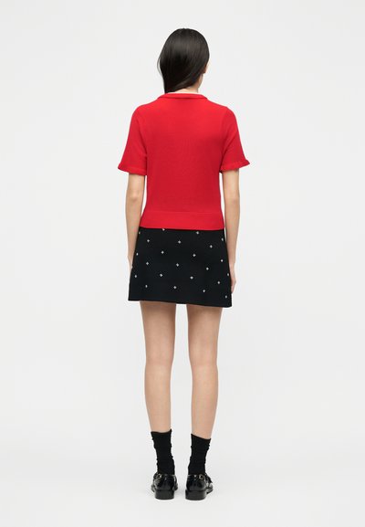 Γυναίκα που στέκεται γυρισμένη με την πλάτη φορώντας κόκκινη κοντομάνικη μπλούζα, μαύρη μίνι φούστα με λευκά μοτίβα σταυρού, μαύρες κάλτσες και μαύρα παπούτσια.