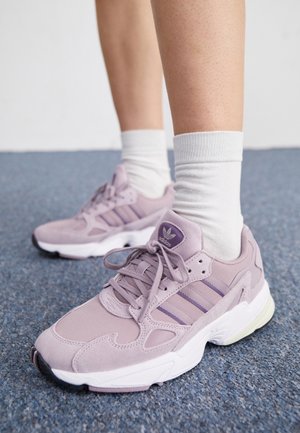 Personne portant des chaussettes blanches et des baskets Adidas violet pastel, debout sur un sol recouvert de moquette bleue.