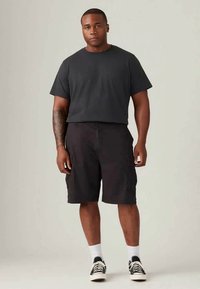 Schwarzes kurzärmliges T-Shirt kombiniert mit knielangen schwarzen Cargo-Shorts. Das Model trägt weiße Socken und schwarze Sneakers mit weißen Akzenten.