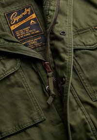 Veste de terrain M65 vert olive avec fermeture éclair en bronze, deux poches avant, coutures détaillées et étiquette en tissu indiquant la marque et des informations de style.