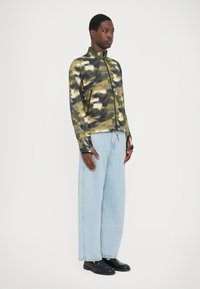 Green and beige camo zip jacket with a high collar, paired with light blue wide-leg jeans and black shoes. Smooth, lightweight fabric.