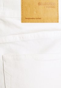 Tejido de mezclilla blanca con dos bolsillos traseros, una etiqueta de cuero con "SELECTED / FEMME" y detalles de costura, que enfatizan un diseño limpio y minimalista.