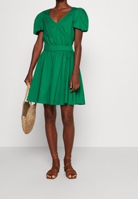 Grön klänning med V-ringning, puffärmar, bälte i midjan och svängd kjol. Matchad med bruna sandaler med remmar och en vävd väska.