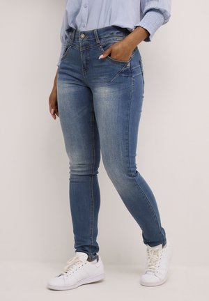 Skinny jeans in medium blue denim, featuring a faded wash, five pockets, and contrasting stitching. Worn with white sneakers.