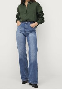 Camicia verde a maniche lunghe con bottoni, abbinata a jeans a vita alta blu a zampa d'elefante. Scarpa nera completa l'outfit.