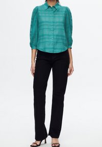 Mujer vistiendo una blusa de botones a cuadros transparente en color verde azulado con mangas abullonadas, pantalones negros de pierna recta y tacones negros abiertos en los dedos.