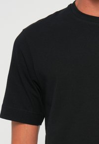 Nahaufnahme einer Person, die ein einfaches schwarzes T-Shirt mit kurzen Ärmeln und Rundhalsausschnitt vor einem hellen Hintergrund trägt.