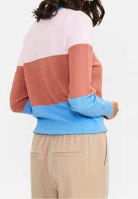 Farbblock-Pullover mit texturierten Bereichen in Pink, Rost und Blau, mit geripptem Saum und Bündchen. Kombiniert mit hellbeigen Hosen.