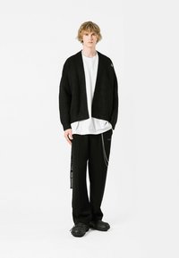 Cárdigan negro texturizado sobre una camisa blanca, combinado con pantalones negros con un acento de cadena y zapatos negros texturizados.