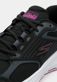 Chaussure de sport noire avec des accents gris, tige en mesh texturé, lacets plats et logo rose. Elle possède une semelle blanche avec un motif.