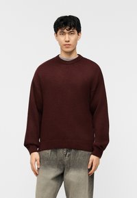 Homme portant un pull en maille bordeaux et un jean gris délavé, debout devant un fond blanc uni.