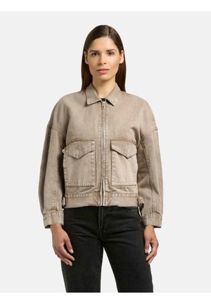 Vrouw draagt een lichtbruine jas met ritssluiting en grote klepzakken aan de voorkant, en zwarte broek, staand tegen een effen witte achtergrond.