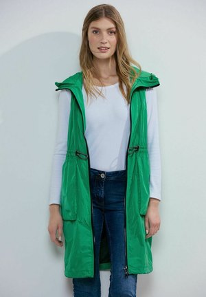 Gilet vert, léger, sans manches avec capuche, deux poches avant, taille à cordon de serrage et fermeture éclair intégrale, porté sur un top blanc et un jean.