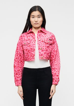 Frau trägt eine kurze rosa Jacke mit Leopardenmuster über einem weißen Oberteil und schwarzen Hosen, steht vor einem einfarbigen hellen Hintergrund.